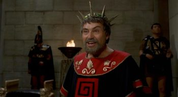 Un homme barbu, qui doit être roi, affiche un visage satisfait, voire rieur ; il porte une couronne aux piques proéminente et est filmé à hauteur de taille ; il est vêtu d'une robe de prestige, au col rouge muni de symboles d'or brodés ; derrière lui, brûle un brasero contre un mur, et l'on aperçoit deux gardes antiques ; l'un des deux gardes est muni d'un casque ; plan du film LES TITANS, réédité par Rimini Editions.