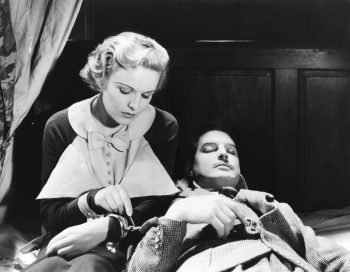 Une jeune femme blonde est assise menottée à un homme dormant à ses côtés ; elle est concentrée sur son poignet, tentant patiemment de déverrouiller les menottes à l'aide d'une épingle ou d'un autre objet de ce genre ; l'homme est Robert Donat, couché et endormi, en costume ; la femme, jouée par Madeleine Carroll, porte un haut élégant, majoritairement blanc, accompagné d'un joli nœud papillon ; plan du film LES 39 MARCHES, réédité par Elephant Films