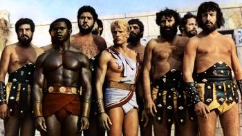 Une dizaine d'esclaves ou de gladiateurs sont filmés à mi-cuisse et regardent tous dans la même direction ; tous sont des hommes, tous blancs, à l'exception d'un homme noir ; tous ont le torse nu et porte un pagne antique ornementé de cuir ou de fer ; au milieu d'eux se détache le héros, un homme blond vêtu d'un pagne blanc et d'un vêtement déchiré (blanc également) qui lui ceint pour partie le torse ; derrière eux, on devine les pierres blanches d'un haut mur ou de remparts ; plan du film LES TITANS, réédité par Rimini Editions.