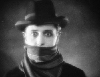 Le visage du "lodger", le locataire du film muet de Hitchcock, joué par la vedette Ivor Novello ; l'homme porte un chapeau noir et a la bouche recouverte d'une écharpe également enroulée autour de son cou ; il a le teint pâle, tranchant avec ses vêtements sombres, et écarquille grand les yeux ; plan du film LES CHEVEUX D'OR (THE LODGER), réédité par Elephant Films.