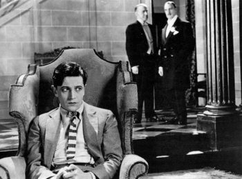 Un jeune homme (joué par Ivor Novello) en costume et portant une cravate rayée est assis inquiet à l'avant-plan sur un très beau fauteuil ; derrière lui, on aperçoit deux hommes en costume sombre en train de discuter ; il doit s'agir du père du jeune homme ainsi que d'un domestique ; la pièce témoigne d'un intérieur luxueux, on y aperçoit, sur la droite du plan, une colonne de style antique, tandis que le sol est un beau carrelage ; plan du film C'EST LA VIE (DOWNHILL), réédité par Elephant Films.