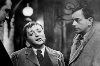 Un homme au front fendu d'une cicatrice fume une cigarette, le visage penché et l'air un peu hautain, aux côtés de deux autres personnages ; l'homme en question est joué par Peter Lorre, il a les cheveux courts et porte un costume cravate ; à la gauche du plan, on aperçoit les traits peu distincts d'un autre homme ; celui situé à la droite du plan est bien plus visible, il porte un manteau gris clair, une cravate et regarde les deux autres individus d'un air interrogateur ; derrière eux, on devine un plafonnier allumé et des décors en fer forgé ; plan du film L'HOMME QUI EN SAVAIT TROP (1934), réédité par Elephant Films.