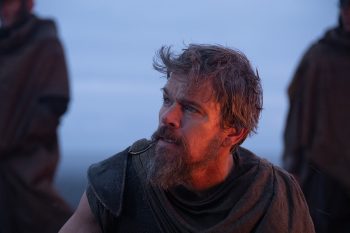 Ulysse, joué par Matt Damon, est filmé en plan rapproché ; il porte une barbe et les cheveux ébouriffés, vêtu d'une simple toge de couleur sombre ; à ses côtés, on devine la présence de deux individus debout, tandis que le bleu du ciel et de la mer domine à l'arrière-plan ; plan du film L'ODYSSEE de Christopher Nolan.
