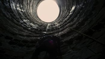 La sortie circulaire d'un puits filmé en contre-plongée ; les pans de l'intérieur du puits sont particulièrement sombres, tandis qu'un cercle lumineux pointe dans le haut de l'image ; il s'agit de l'entrée ou de la sortie du puits ; une chaîne semble relier le trou du puits au sol en bas ; tout est extrêmement sombre et ténébreux, à l'exception de ce cercle de lumière ; plan du film THE DARK KNIGHT RISES de Christopher Nolan.