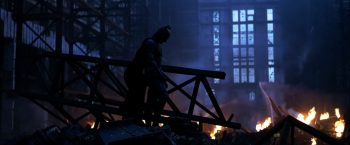 Batman se tient debout, la tête penchée vers le sol, au beau milieu de décombres encore en feu ; il occupe le centre du plan telle une ombre, tandis qu'on aperçoit des débris et du feu au sol ; une fenêtre, à l'arrière-plan, éclaire faiblement cette image sombre ; plan du film THE DARK KNIGHT : LE CHEVALIER NOIR de Christopher Nolan.
