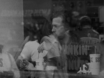 Sur une image en noir et blanc, un homme apparaît de l'autre côté d'une vitre en train de touiller dans sa tasse de café en carton ; il est assis à une table ou à un comptoir et à le visage relevé en train d'observer vers la gauche du plan ; on aperçoit la silhouette d'autres clients et d'autres boissons ; plan du film FOLLOWING, LE SUIVEUR de Christopher Nolan.