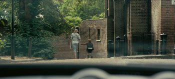 Une femme et un enfant se tiennent la main, de dos, et au loin ; tous deux sont observés depuis l'habitacle d'une voiture, de l'autre côté du parebrise ; les deux personnages avancent vers des bâtiments en briques bordés d'arbres et de végétation ; plan du film TENET de Christopher Nolan.