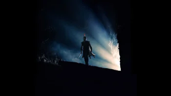 Un héros grec, qui doit être Ulysse, est filmé de nuit, au loin, en contre-plongée ; il a l'air de longer le bord d'une falaise et un halo lumineux perce derrière lui, accompagnant sa progression nocturne ; plan du film L'ODYSSEE de Christopher Nolan.