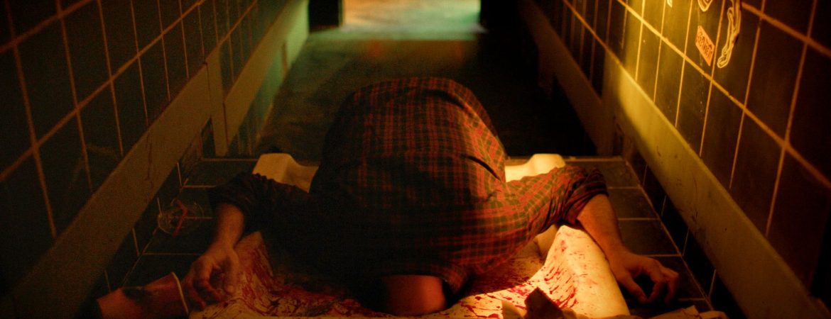 Un homme coincé dans les toilettes turcs d'un bar ; on ne voit dépasser que ses épaules et ses bras ; plan issu du film Flush de Gregory Morin.