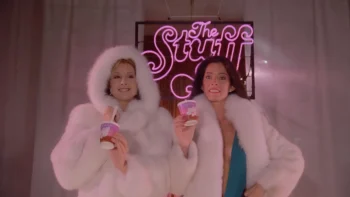 Deux femmes emmitouflées dans des manteaux blancs duveteux et posant devant un néon rose très seventies présentent un petit pot rose de ce qui semble être une crème glacée ; le néon rose indique "The Stuff" ; la femme sur la gauche est blonde et a rabattu le capuchon duveteux de son manteau ; celle de droite est brune et a de longs cheveux lâchés ; plan du film THE STUFF, réédité par Rimini.