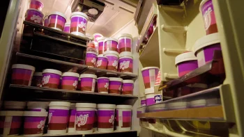 Des rayons de magasins remplis de pots bariolés de rose, de mauve et de brun contenant le produit "The Stuff" ; une lumière d'hôpital éclaire ces rayons probablement frigorifiés ; plan du film THE STUFF, réédité par RIMINI.