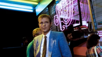 Le personnage de Mo dans le film "The Stuff" de Larry Cohen filmé à hauteur de taille ; il a le visage passablement inquiet et porte un veston gris, une chemise blanche et une cravate rouge ; derrière lui, on devine trois autres personnages ; la pièce, qui pourrait être un restaurant ou un autre établissement commercial, est éclairée par des néons cliniques au plafond, tandis que sur une vitre, à droite du plan, apparaît le néon rose "The Stuff" ; plan du film THE STUFF, réédité par Rimini.