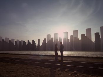 Deux silhouettes lointaines (un homme et une femme) se tiennent face-à-face sur la grève à contre-jour ; derrière la mer placide apparaissent de grands buildings ; le ciel est bleu, émaillé de nuages sans danger, et le soleil rayonne derrière les gratte-ciels ; plan du film INCEPTION de Christopher Nolan.
