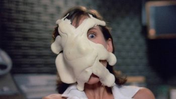 Une matière blanche squammeuse semble attaquer le visage d'une femme apeurée ; la matière blanche adhère au visage de la femme, en le recouvrant quasi en intégralité, à l'exception d'un de ses yeux ; la femme est brun et son faciès est filmé en plan rapproché ; on devine qu'elle porte un haut ou une robe blanche ; plan du film THE STUFF, réédité par Rimini.