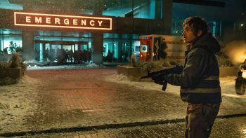 Sous la neige, un jeune homme armé se tient devant les urgences d’un hôpital, une veste sombre à bandes réfléchissantes sur le dos ; le fusil tenu à deux mains, il regarde avec inquiétude vers la gauche, hors champ ; derrière lui, une ambulance et plusieurs silhouettes apparaissent devant l’entrée vitrée, dominée par l’enseigne rouge « EMERGENCY » ; plan du film RESIDENT EVIL de Zach Cregger.