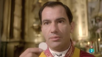 En gros plan, le visage d'un prêtre en habits liturgiques rouges et dorés, qui tient devant lui une hostie blanche ; le regard fixe, le visage grave, il semble absorbé par sa contemplation ; derrière lui, on devine l'arrière de l'autel d'une église, aux décorations dorées mais floues ; plan du film LE PRÊTRE, réédité par Artus Films.