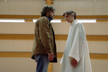 the-holy-boy-strippoli-sergio-matteo-critique-2026 Dans une grande salle lumineuse aux poutres de bois apparentes, un homme barbu en veste marron fait face à un jeune homme très mince, entièrement drapé dans une longue tunique blanche ; le premier le regarde gravement, légèrement voûté ; le second, les bras dissimulés sous l’étoffe ample, incline la tête et lui répond par un regard calme et impassible ; filmés en légère contre-plongée, les deux hommes restent immobiles, séparés par un faible espace qui donne à leur face-à-face une tension presque solennelle ; plan du film THE HOLY BOY de Paolo Strippoli.