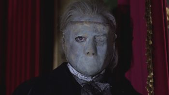 En gros plan, une silhouette aux cheveux gris apparaît entre deux lourds rideaux rouges ; son visage est dissimulé sous un masque gris presque dépourvu de traits, sans bouche ; un seul œil apparaît à travers une ouverture ; l’autre est recouvert par le masque et marqué d’une cicatrice ; vêtue de noir, la silhouette regarde sur le côté ; plan du film LE FANTOME DE L'OPERA de Terence Fisher.