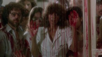 Une foule se presse derrière une vitre ruisselante de sang ; au centre, une femme brune en chemise blanche plaque ses deux mains ensanglantées contre le verre, tandis que de longues traînées rouges coulent devant son visage ; autour d’elle, plusieurs personnes couvertes de sang regardent vers l’extérieur, leurs visages partiellement brouillés par la vitre maculée ; plan du film PIRANHA 2 de James Cameron.
