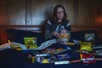 Assise au centre d’un grand lit, une jeune femme brune à lunettes regarde droit devant elle, les jambes repliées et une main plongée dans un pot de pop-corn ; le lit qui l’entoure est entièrement jonché de sachets de chips, bonbons, chocolats et autres friandises aux emballages colorés; derrière elle, les murs lambrissés de bois baignent dans une lumière sombre et bleutée, réchauffée sur la droite par une lueur orangée.