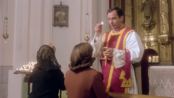 Devant l’autel doré d’une église, le prêtre tient un calice et élève une hostie ; deux femmes agenouillées devant lui attendent de communier ; l’une est vêtue de noir et porte un voile ; plan du film LE PRÊTRE, réédité par Artus Films.