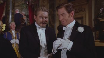 Au milieu d’une réception élégante, deux hommes en smoking noir et nœud papillon blanc se tiennent côte à côte ; à droite, un homme brun, une fleur blanche à la boutonnière, ajuste ses gants avec un léger sourire ; à gauche, un homme blond et barbu l’observe de côté, le visage sérieux ; derrière eux, des invités en tenue de soirée se pressent dans le décor fastueux du théâtre ; plan du film LE FANTOME DE L'OPERA de Terence Fisher.