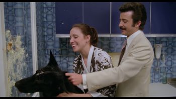 la-créature-couple-cuisine-eloy-iglesia-artus-2026 Dans une cuisine aux murs carrelés de bleu, une femme rit tandis qu’un homme moustachu, en costume clair, tient près d’elle un grand chien noir ; tous trois sont tournés vers un mur couvert de nourriture écrasée ; plan du film LA CREATURE, réédité par Artus Films.