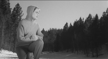 Une femme en pull à capuche sur le bord gauche de l'image ; regardant au loin, dans un paysage forestier rempli de sapins ; plan issu du film la bête de la caverne hantée de Monte Hellman.