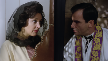 De part et d’autre de la cloison d’un confessionnal, une femme au voile noir et le prêtre, vêtu d’une étole violette et or, se regardent en parlant à voix basse ; plan du film LE PRÊTRE, réédité par Artus Films.