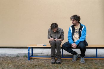 the-holy-boy-strippoli-sergio-matteo-banc-critique-2026 Assis côte à côte sur un banc devant un immense mur beige, un jeune homme en chemise à carreaux, tête baissée, s’affaire minutieusement sur un petit objet qu’il tient entre ses doigts ; à sa droite, un homme aux cheveux gris et bouclés, en survêtement bleu et noir, l’observe en silence, les mains jointes ; le vaste mur nu qui les surplombe accentue leur isolement dans le cadre ; plan du film THE HOLY BOY de Paolo Strippoli.