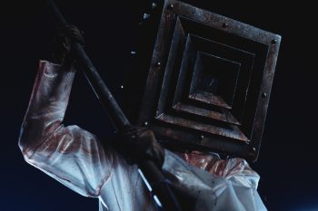Dans l’obscurité, une silhouette en combinaison blanche éclaboussée de sang brandit une longue arme à deux mains ; son visage est entièrement dissimulé sous un lourd masque métallique carré, composé de cadres concentriques autour d’une ouverture noire ; filmée en forte contre-plongée et éclairée d’une lumière froide, elle se détache sur un fond presque entièrement noir.