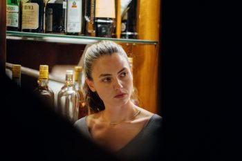 the-holy-boy-strippoli-michela-vergano-bar-critique-2026 Dans un bar, une jeune femme brune aux cheveux attachés se tient devant des rangées de bouteilles ; encadrée au premier plan par deux silhouettes floues, elle tourne la tête vers l’une d’elles et lui adresse un regard attentif et sérieux ; plan du film THE HOLY BOY de Paolo Strippoli.