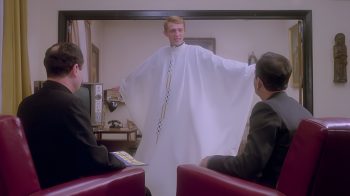 Dans un salon, un jeune prêtre en aube blanche se tient debout, les bras largement écartés, face à deux prêtres assis dans des fauteuils rouges ; il semble leur présenter ou essayer son nouvel habit liturgique ; plan du film LE PRÊTRE, réédité par Artus Films.