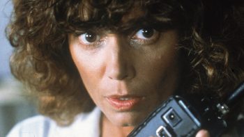 En très gros plan, une femme brune aux cheveux bouclés tient un téléphone noir contre sa joue ; les yeux écarquillés, elle fixe quelque chose hors champ, les lèvres entrouvertes ; une moitié de son visage est vivement éclairée tandis que l’autre reste plongée dans l’ombre ; plan du film PIRANHA 2 de James Cameron.