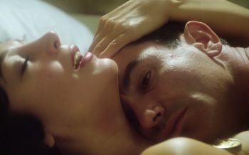 En gros plan, le prêtre repose la tête contre la poitrine nue d’une femme ; elle garde les yeux fermés et pose une main sur ses cheveux, tandis que lui fixe silencieusement devant lui ; plan du film LE PRÊTRE, réédité par Artus Films.
