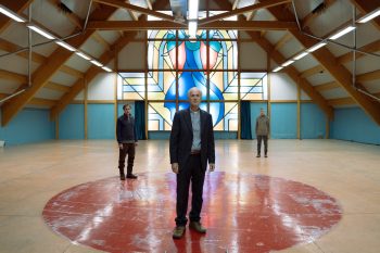 the-holy-boy-strippoli-pretre-salle-chapelle-critique-2026 Dans une immense salle aux poutres de bois, trois hommes se tiennent immobiles et très éloignés les uns des autres ; au premier plan, un homme âgé en costume sombre se dresse au centre d’un large cercle rouge peint au sol ; deux hommes plus jeunes restent en retrait, de part et d’autre ; derrière eux, un gigantesque vitrail abstrait bleu, rouge et jaune domine la salle ; la composition parfaitement frontale et symétrique place l’homme âgé au centre de l’espace ; plan du film THE HOLY BOY de Paolo Strippoli.