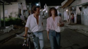 De nuit, un homme et une femme marchent côte à côte dans une rue presque déserte ; chemise blanche ouverte, l’homme pousse son vélo et tourne le visage vers sa compagne ; en chemisier rose et jean, un sac blanc à l’épaule, elle continue de regarder droit devant elle ; autour d’eux, les petites maisons sont faiblement éclairées ; plan du film PIRANHA 2 de James Cameron.