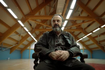 the-holy-boy-strippoli-père-mauro-critique-2026 Seul au milieu d'une vaste, un homme barbu aux cheveux grisonnants est assis dans un fauteuil en bois, les mains jointes sur le ventre. ; il lève les yeux sur le côté, le visage fermé ; filmé en contre-plongée, il se détache sous l’enchevêtrement symétrique des poutres et des néons du plafond ; plan du film THE HOLY BOY de Paolo Strippoli.