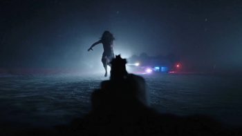 Dans une rue nocturne envahie de brume, une arme à feu, vue dans l’axe du tireur, pointe vers une silhouette qui court droit vers lui ; éclairée à contre-jour par de puissants phares, celle-ci apparaît presque entièrement noire, les bras écartés ; au loin, des gyrophares bleus et rouges percent le brouillard ; plan du film RESIDENT EVIL de Zach Cregger.