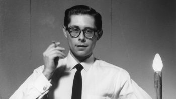 Photo en noir et blanc de Roland Loomis ; aka Fakir Musafar ; avec des lunettes habillé d'une chemise blanche et d'une cravate noire ; fumant une cigarette dans sa main droite ; positionné à gauche d'une fausse chandelle ; photo issu du documentaire a body to live in de Angelo Madsen.