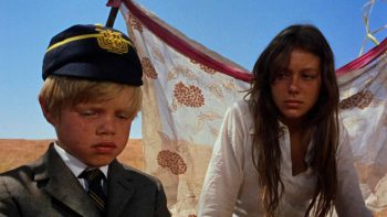 Une jeune fille brune , cheveux bruns et longs ; fatiguée ; regardant par sa droite son petit frère ; blond en costume avec une casquette ; dans le désert ; protégés par un voile blanc derrière eux ; plan issu de Walkabout de Nicolas Roeg.