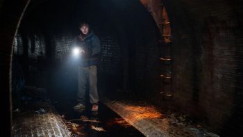Seul dans un tunnel voûté de briques, un jeune homme armé éclaire l’obscurité avec une lampe fixée à son arme ; la bouche entrouverte, il regarde devant lui ; le sol mouillé et couvert de détritus reflète son faisceau, tandis qu’une échelle métallique grimpe le long du mur vers une ouverture faiblement éclairée d’orange ; plan du film RESIDENT EVIL de Zach Cregger.