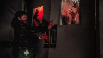 Dans un couloir obscur, un homme vêtu de noir avance une main tendue devant lui, retenant une porte à double battant ; à travers deux petites fenêtres éclairées de rouge, des silhouettes pressent leurs mains ouvertes contre les vitres de la porte ; l’homme tourne le visage vers elles, grimaçant ; à sa taille pend une trousse de secours verte marquée d’une croix blanche ; plan du film RESIDENT EVIL de Zach Cregger.