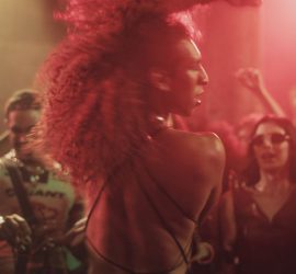 A Margem do Rio / The Riverbank : une femme danse au milieu d’une foule sous des lumières rouges.