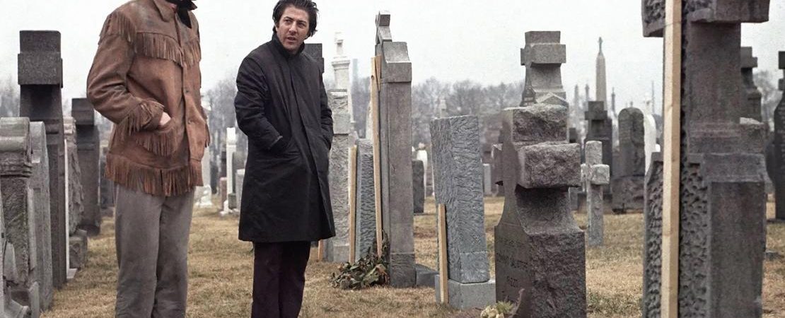 Un homme blond habillé en cow-boy à côté d'un homme brun plus petit avec un manteau ; devant une tombe dans un cimetière ; plan issu du film Macadam cow-boy de John Schlesinger.
