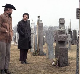 Un homme blond habillé en cow-boy à côté d'un homme brun plus petit avec un manteau ; devant une tombe dans un cimetière ; plan issu du film Macadam cow-boy de John Schlesinger.
