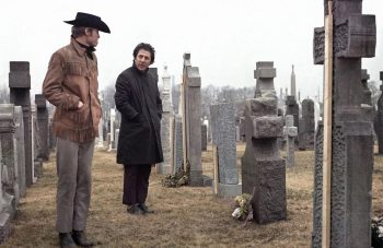 Un homme blond habillé en cow-boy à côté d'un homme brun plus petit avec un manteau ; devant une tombe dans un cimetière ; plan issu du film Macadam cow-boy de John Schlesinger.