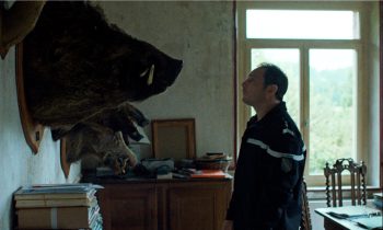 Gendarme de profil regardant la tête d'un énorme sanglier empaillé accroché au mur dans une vieille maison ; plan issu du film l'espèce explosive de Sarah Arnold.
