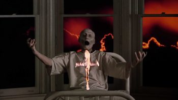 Image cauchemardesque d'un prêtre chauve au visage blanc ; la bouche déformé par un cri ; levant les bras face à une croix dans un décor apocalyptique ; plan issu du film Au-delà du réel de Ken Russel.