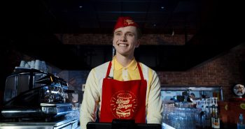 marion-le-corroller-sanguine-fastfood-entretien-2026 Derrière le comptoir d'un restaurant de restauration rapide, un jeune employé blond, vêtu d'une chemise jaune et d'un tablier rouge portant le logo « Bloody Burger », affiche un immense sourire, les yeux écarquillés ; il porte une petite toque rouge assortie et un écouteur à fil torsadé ; de part et d'autre de lui, une machine à café et des rangées de verres occupent le comptoir, dans un intérieur sombre aux murs de briques ; plan du film SANGUINE de Marion Le Corroller.